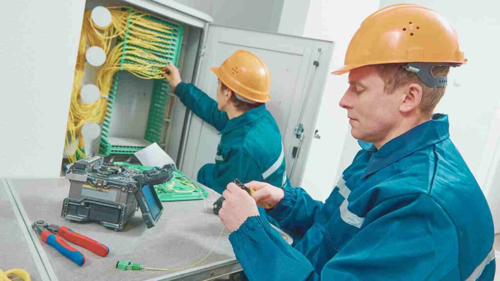 Two men collaborating on fiber optics installation in a well-lit room, focused on their task and surrounded by tools.